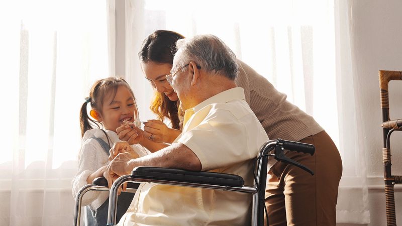 Hospice Volunteer. An elderly man, his daughter and granddaughter smile together in a well-lit room.