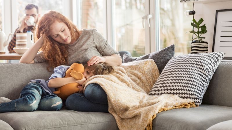 A woman and a little girl cuddled together on a couch. The girl is holding a stuffed animal.