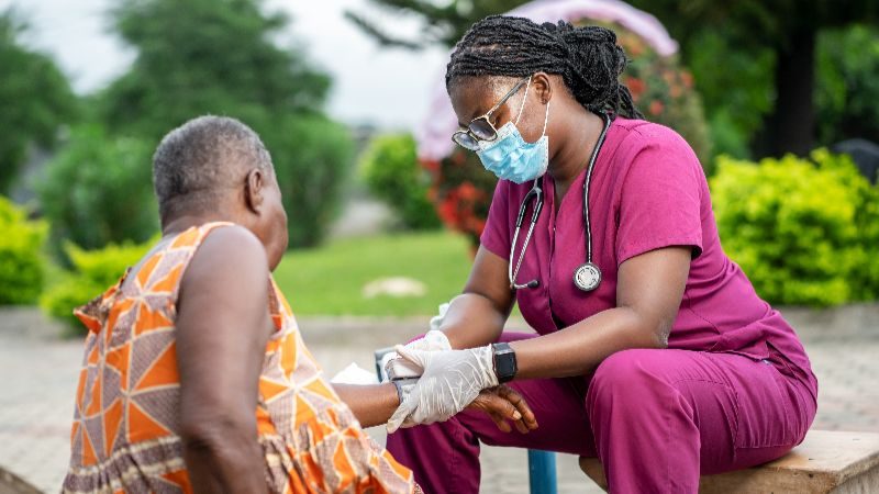 A medical professional sitting outside with an elderly person and providing treatment.