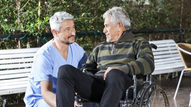 Grey-haired man in blue scrubs kneels by a wheelchair occupied by a man in a green and black striped shirt and black pants