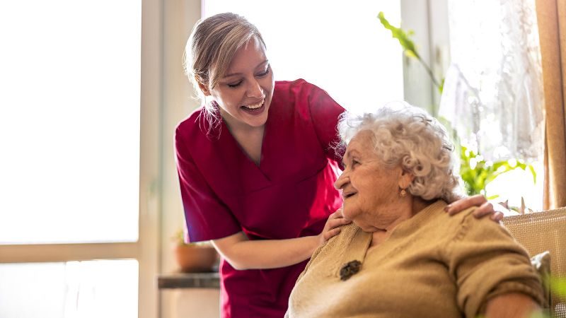 A Nurse And An Elderly Woman Smile At Each Other In The Elderly Woman's Home