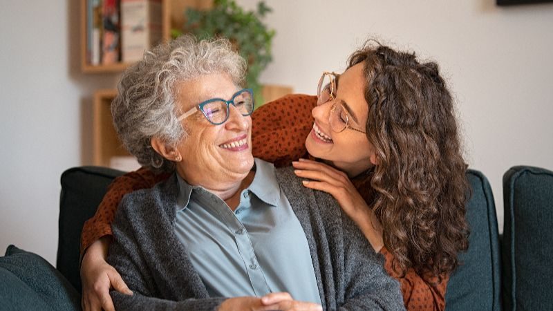 A young woman holding an elderly woman as they smile at each other.